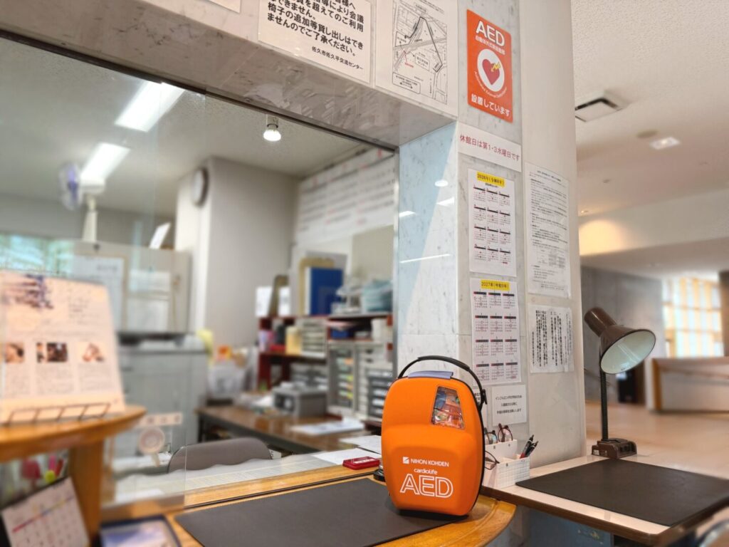 佐久平交流センターの受付窓口に置かれた日本光電製のオレンジ色のAED。柱にAED設置ポスターと休館日の掲示がある