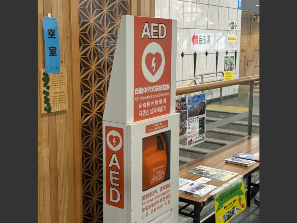 佐久平駅改札内のAED本体アップ。日本光電製のオレンジ色のAEDと赤い案内看板
