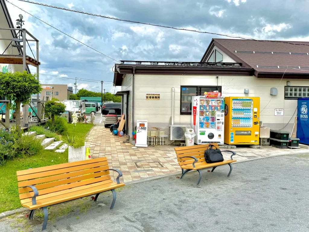市民交流ひろば管理事務所の建物。ベンチと自動販売機が並び、左側に白いAEDスタンドボックスが見える