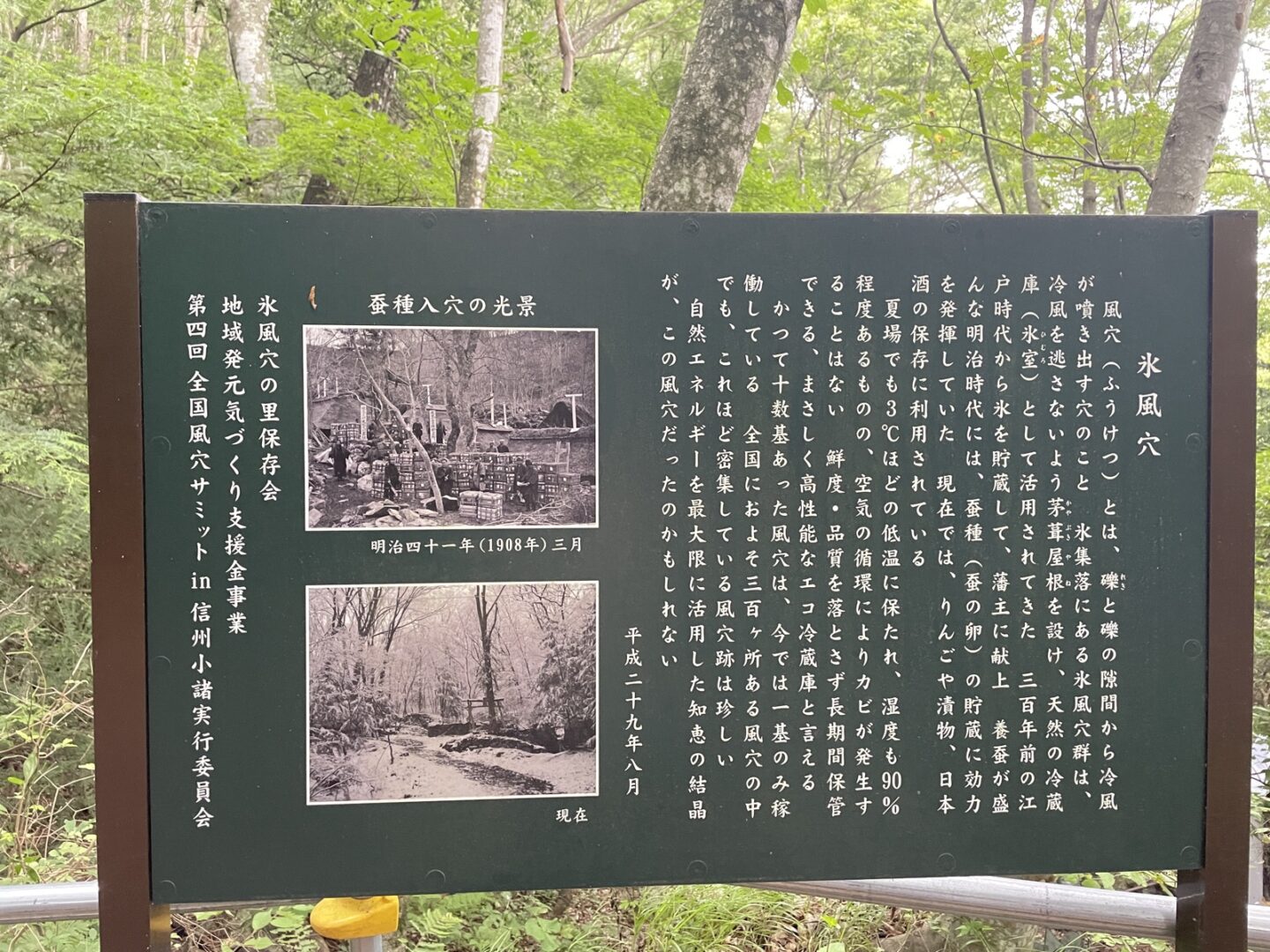 森の中に立つ「氷風穴」の解説看板。明治41年(1908年)の蚕種入穴の光景の古写真と、現在の風穴の写真が並ぶ