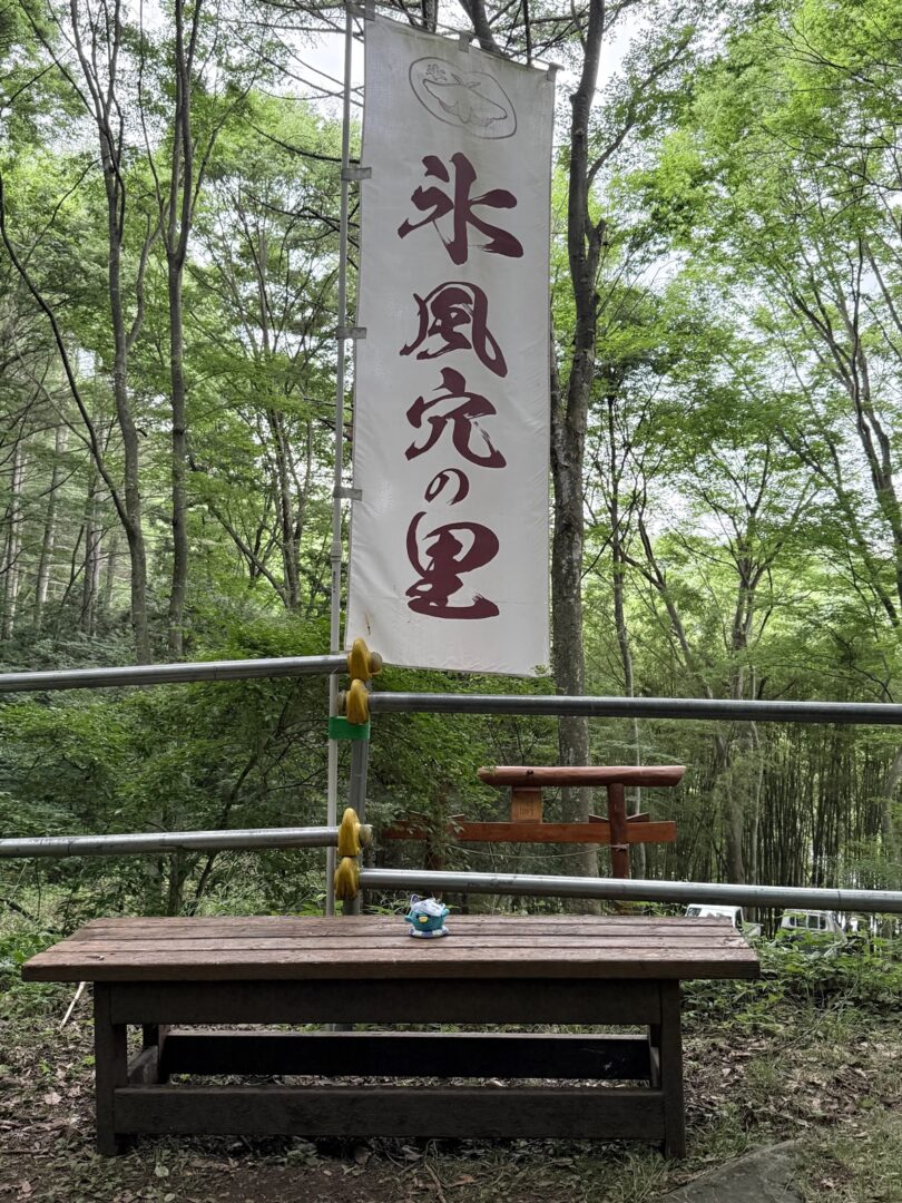 「氷風穴の里」と書かれた白いのぼり旗と、木立の中の小さな鳥居。手前にはベンチとエールくんのマスコット
