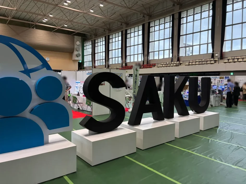 体育館内に置かれた大きな「SAKU」の立体ロゴと、奥に並ぶ展示ブース