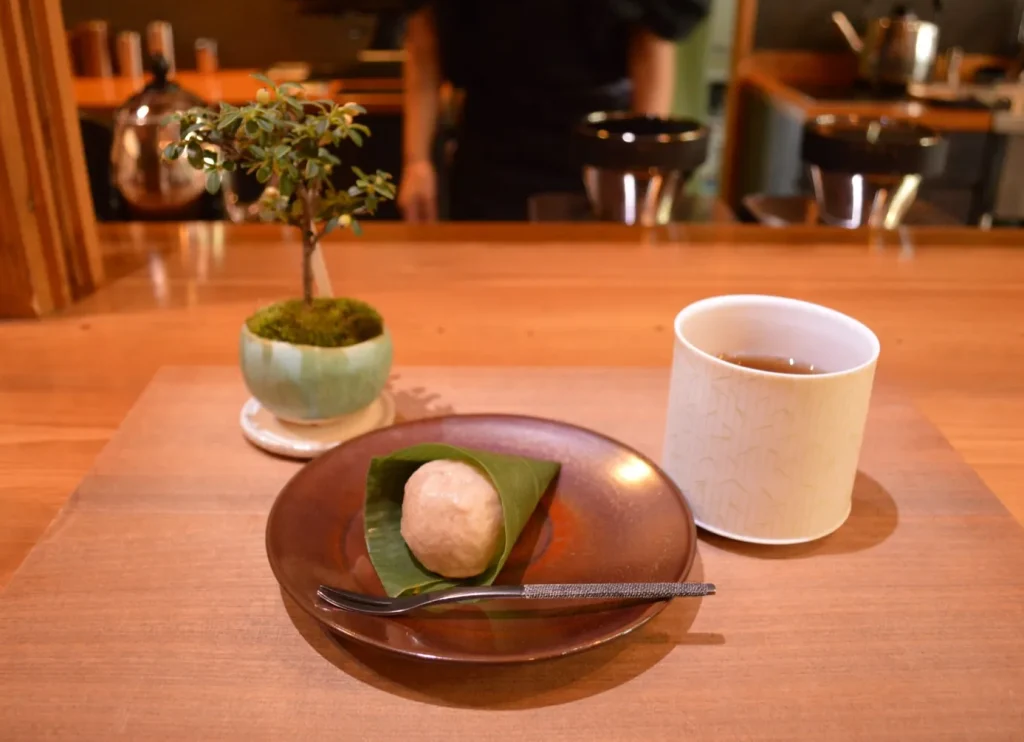 笹の葉に包まれた麩饅頭と和紅茶「梔子」、奥に小さな盆栽